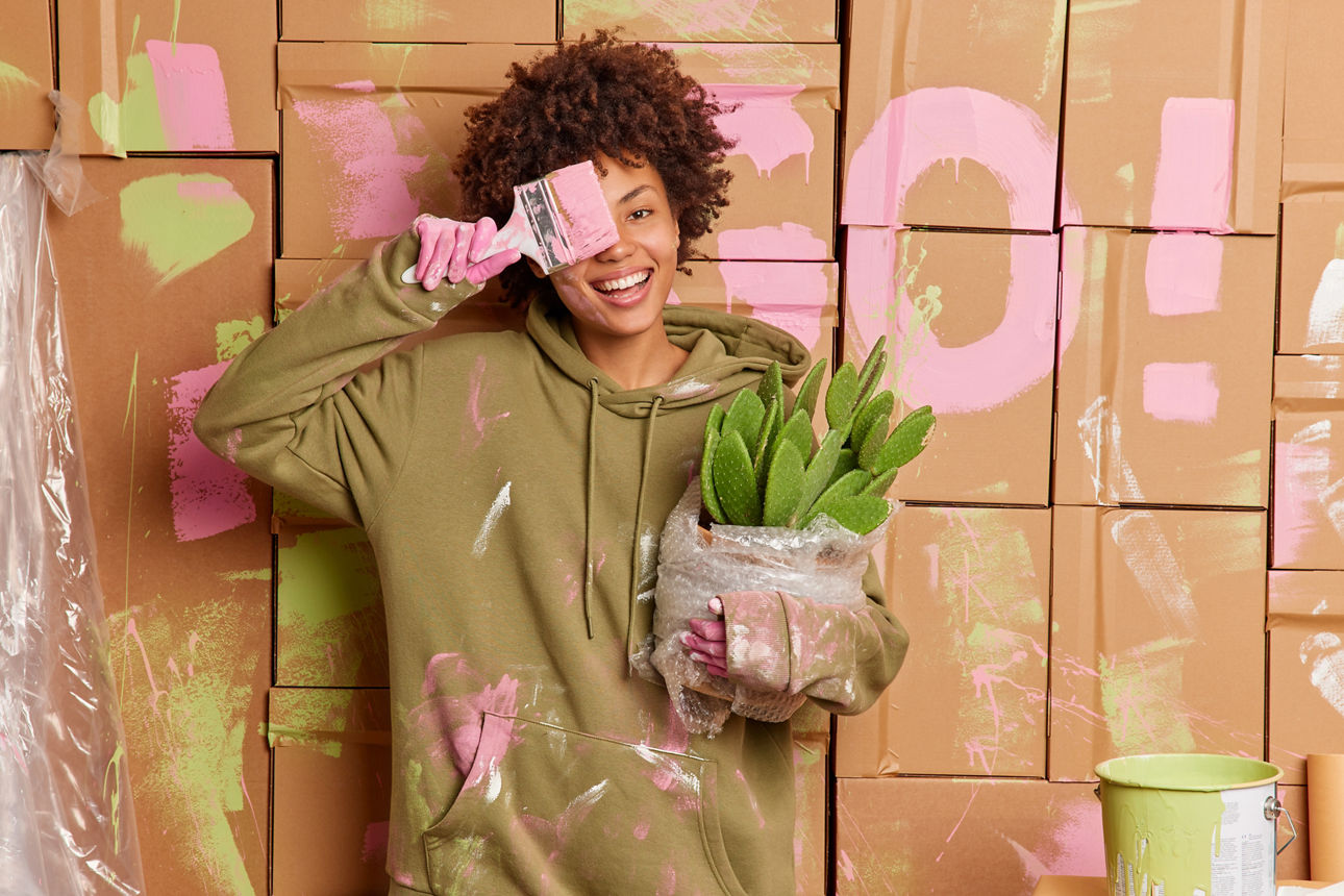 Eine Frau steht mit einem Pinsel in der Hand vor einer Karton-Wand.  Sie lächelt und hält eine Pflanze in der Hand.