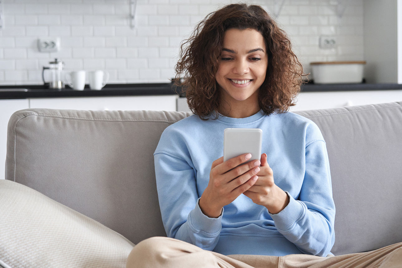 Eine Frau sitzt auf einer Couch und schaut erfreut auf ihr Smartphone