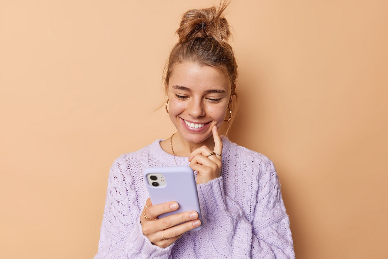 Pleased young woman smiles happily keeps index finger on cheek concentrated into smartphone device reads pleasant message wears casual jumper isolated over beige background. Cellular technology