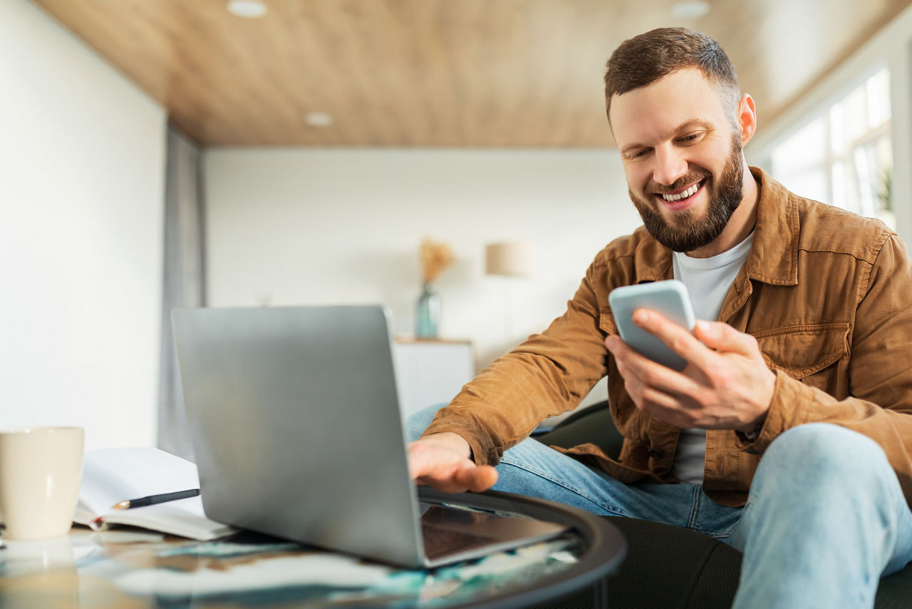 Männlicher Unternehmer mit Smartphone und Laptop, der zu Hause online arbeitet