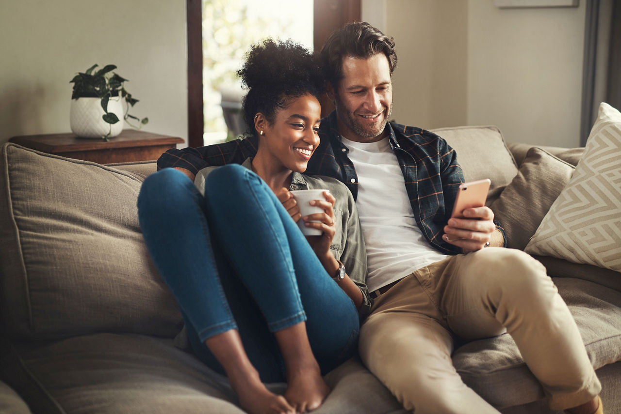 Haftpflichtversicherung Paar liegt kuschelnd und sorglos auf der Couch und schaut auf ein Handy