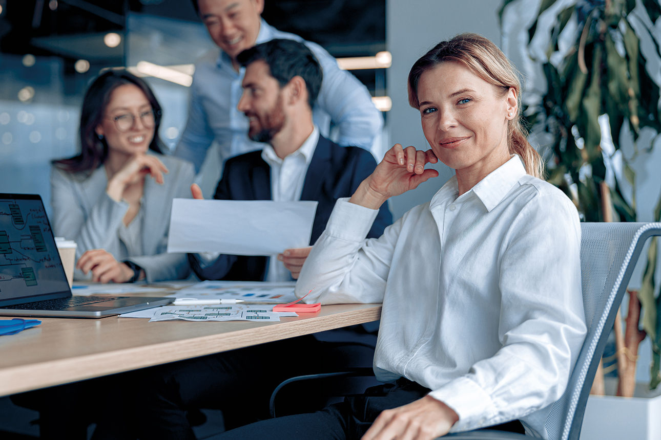 Lächelnde, selbstbewusste Geschäftsfrau sitzt in einer Besprechung im Büro, mit ihren Kollegen im Hintergrund.