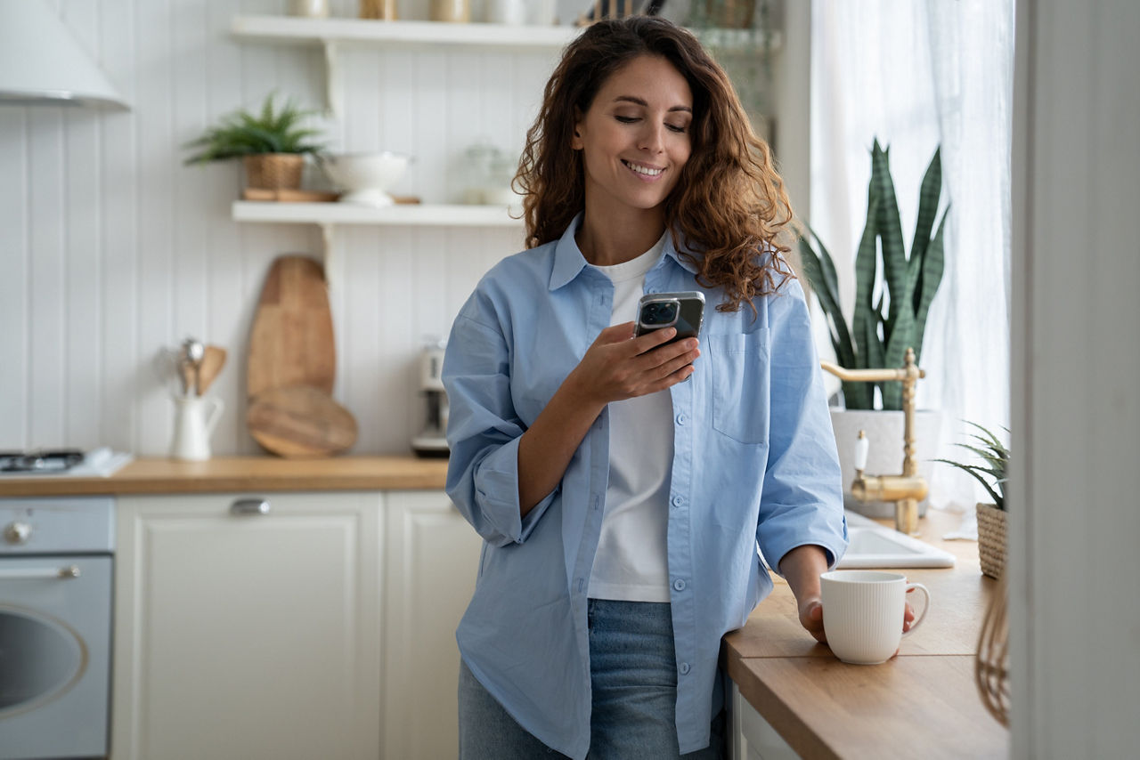 Junge Frau steht in einer hellen Küche am Handy und mit Kaffeetasse in der Hand, sie lächelt zufrieden