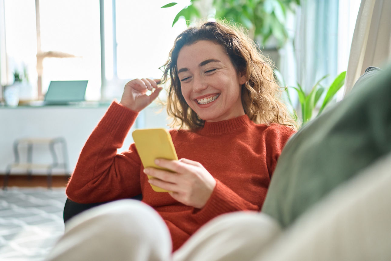 Eine Frau sitzt  lachend auf dem Sofa  und hält ein Smartphone in der Hand 