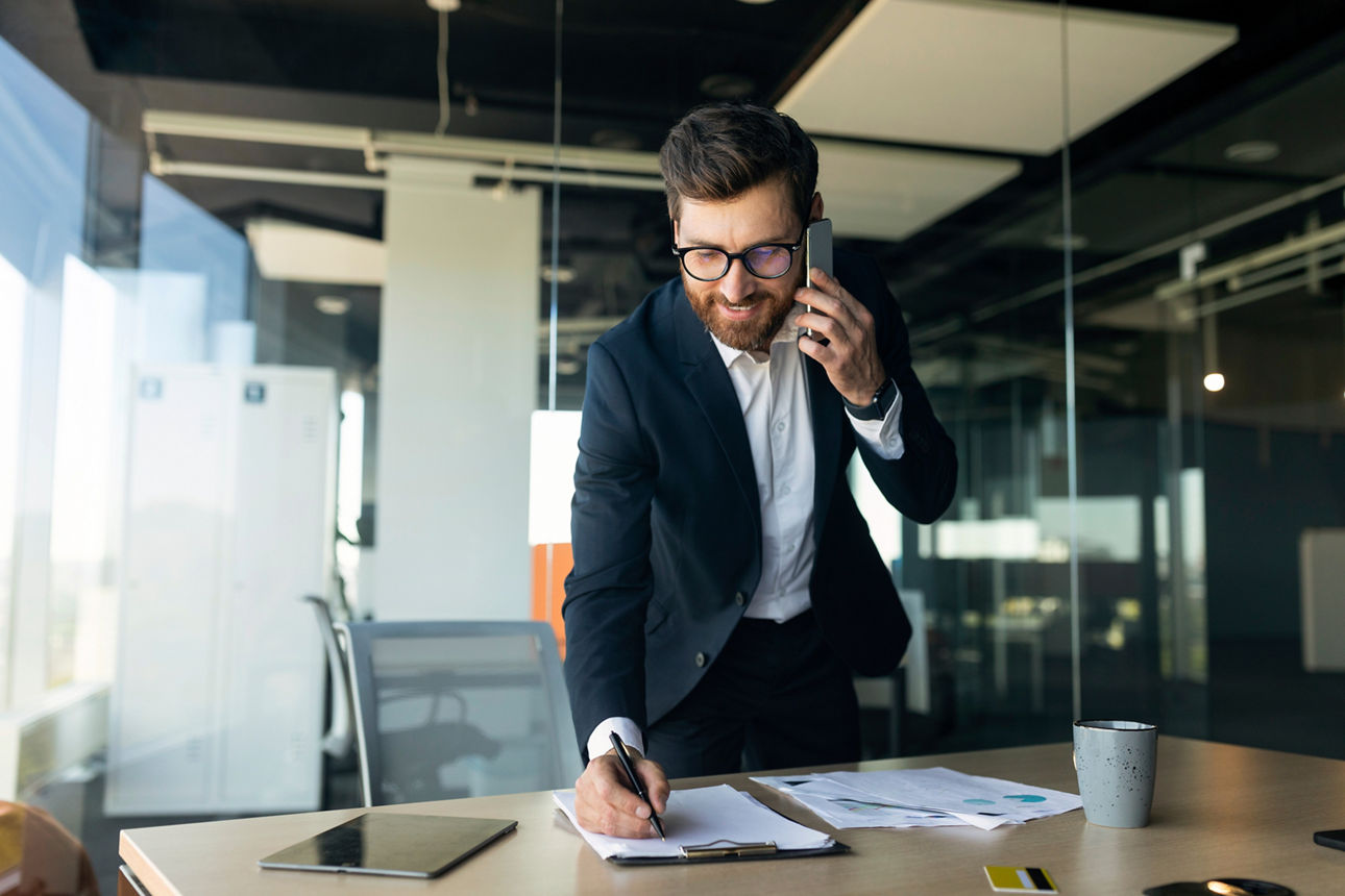 Ein Mann steht mit Handy am Ohr an einem Schreibtisch und macht sich Notizen.