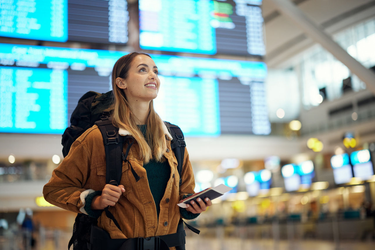 Eine junge Frau steht freudig mit Rucksack und Reisepass am Flughafen.