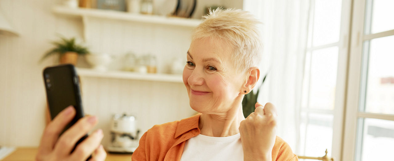 Attractive trendy mature female blogger making funny content standing in kitchen recording stories or making selfie with clenched fist and sly smile. Senior woman using video chat on smartphone