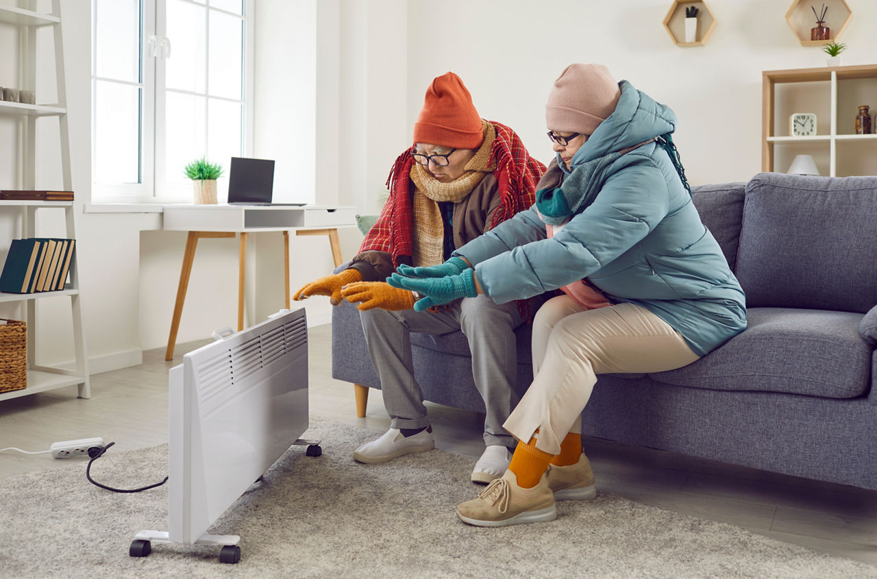 Älteres Paar sitzt zu Hause, gekleidet mit Mütze, Schal und Wintersachen und wärmt sich.