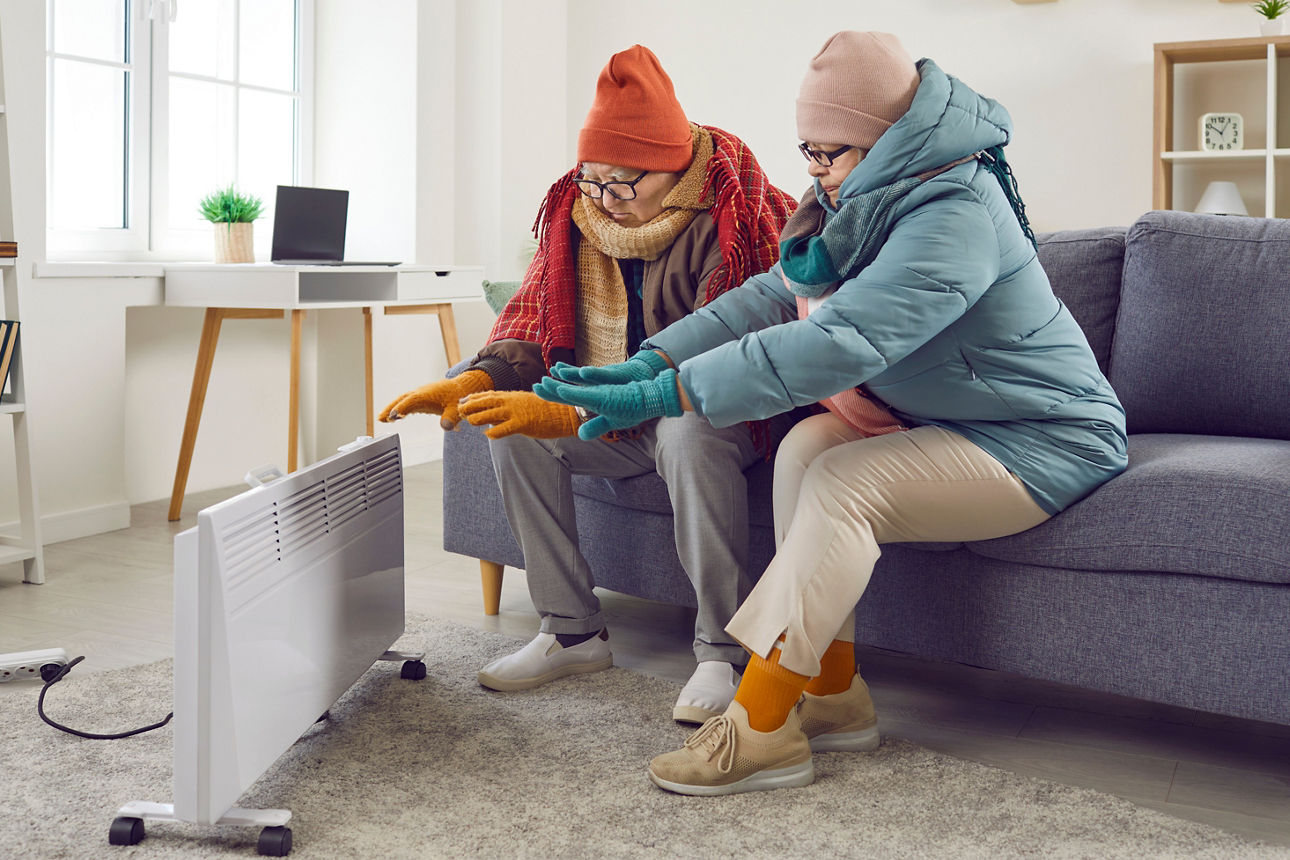 Älteres Paar sitzt zu Hause, gekleidet mit Mütze, Schal und Wintersachen und wärmt sich.