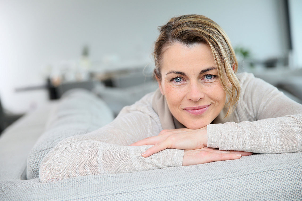 Eine Frau stützt lächelnd ihr Kinn auf ihren verschränkten Händen und sitzt dabei auf dem Sofa.
