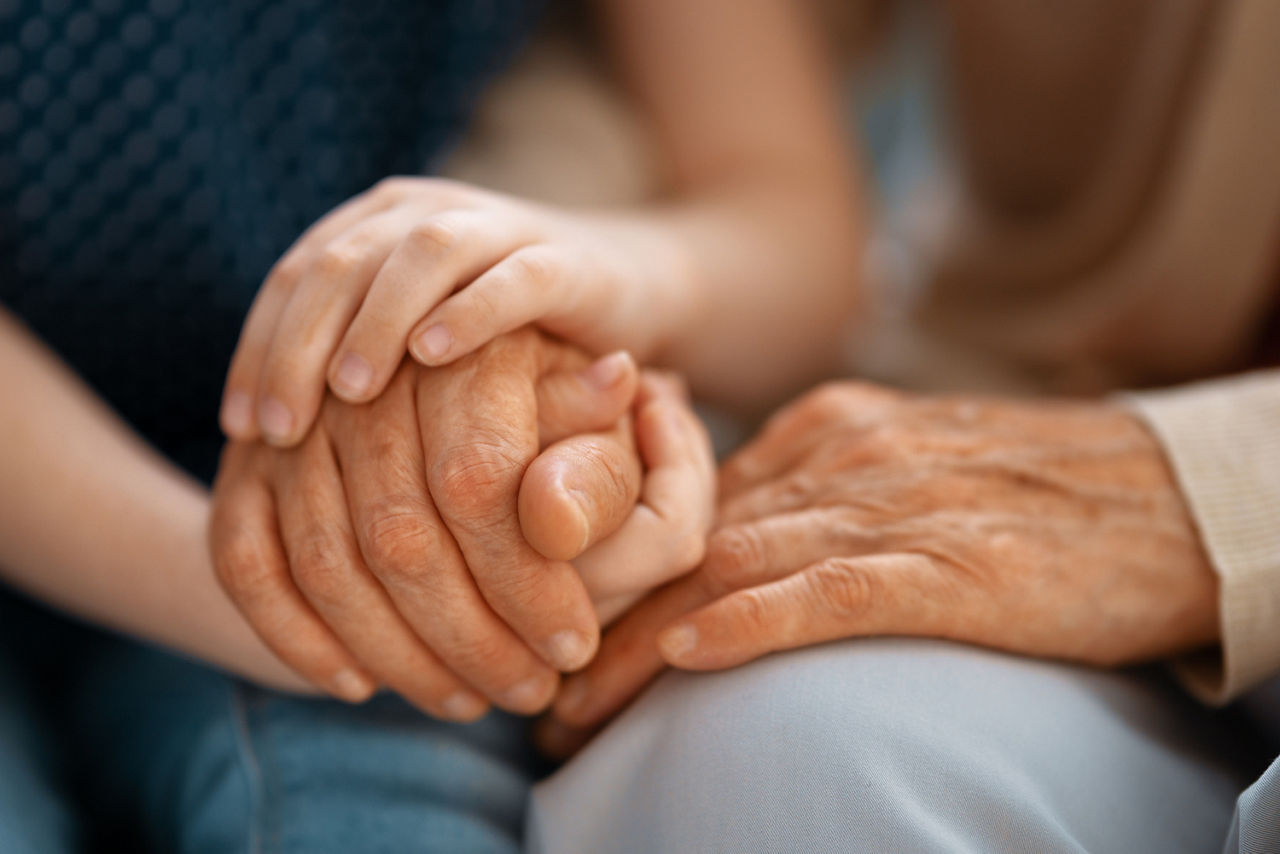 Zwei Menschen halten die Hände ineinander verschlungen. Eine Hand einer jungen Person, eine Hand einer älteren Person.