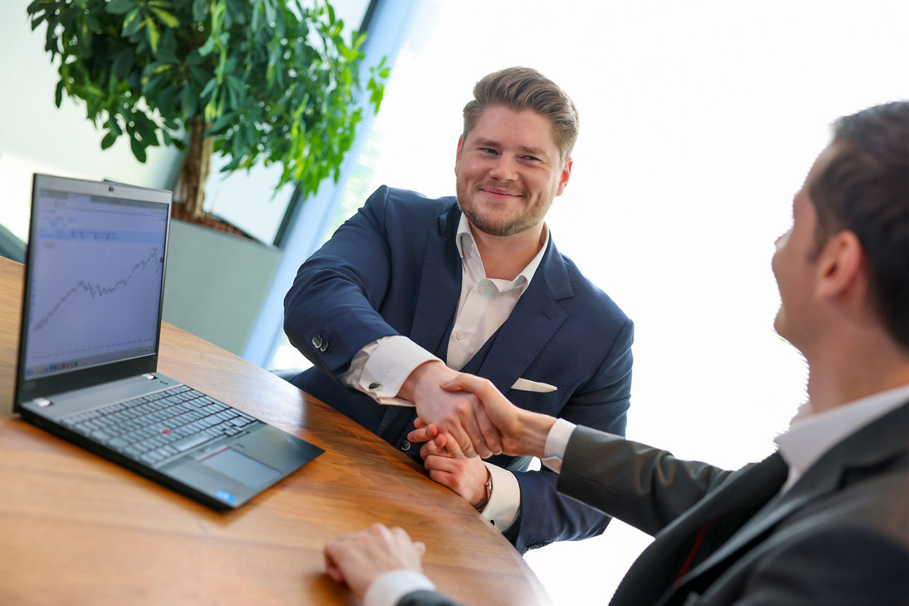 Der Berater Dominik Niers sitzt an einem Tisch auf dem ein Laptop mit Dashboard steht. Er gibt einem Mann die Hand.