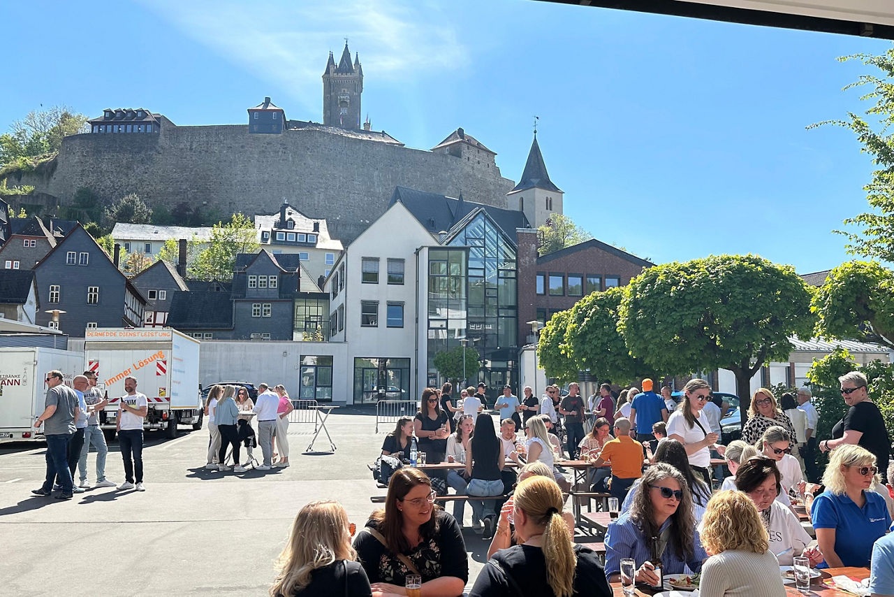 Mitarbeiterinnen und Mitarbeiter der Bank sitzen bei tollem Wetter vor Dillenburger Hauptstelle mit Schloss im Hintergrund.