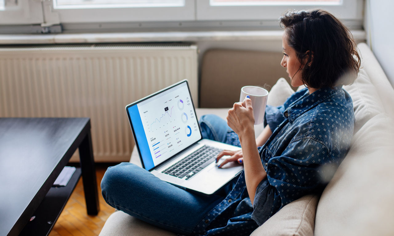 Frau sitzt im Schneidersitz mit einem Laptop auf dem Schoß und einer Kaffeetasse in der Hand auf einem Sofa und schaut sich Aktienwerte an