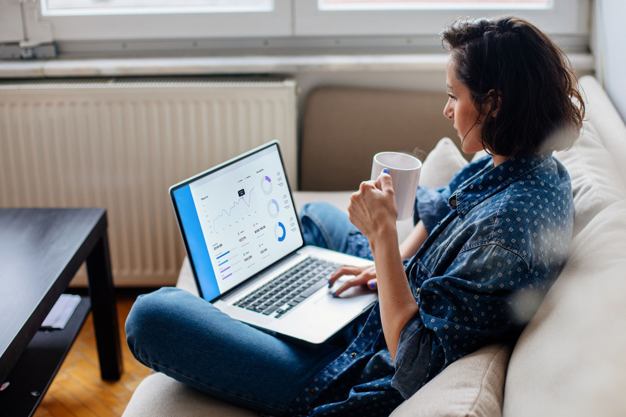 Frau sitzt im Schneidersitz mit einem Laptop auf dem Schoß und einer Kaffeetasse in der Hand auf einem Sofa und schaut sich Aktienwerte an