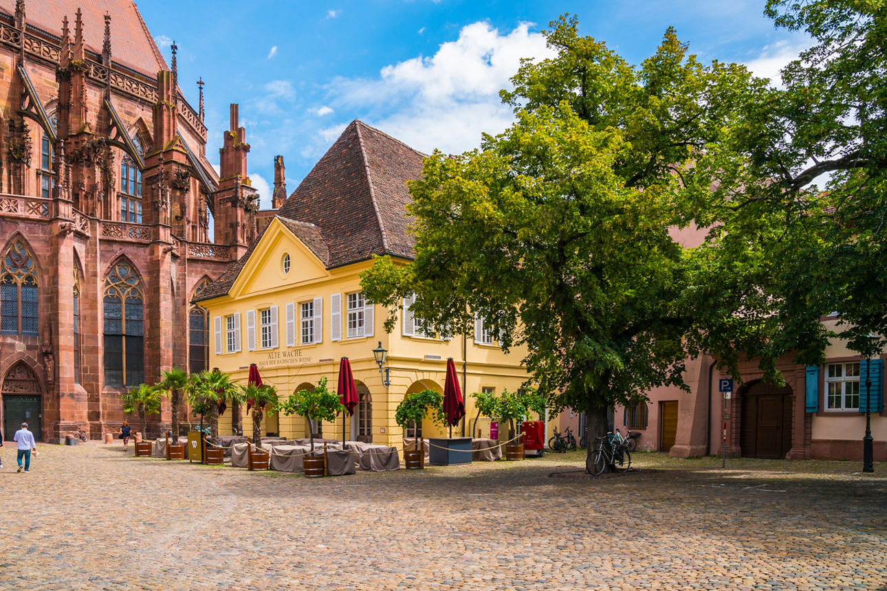 Historischer Platz mit Kirche, gelbem Gebäude und Caféterrasse.