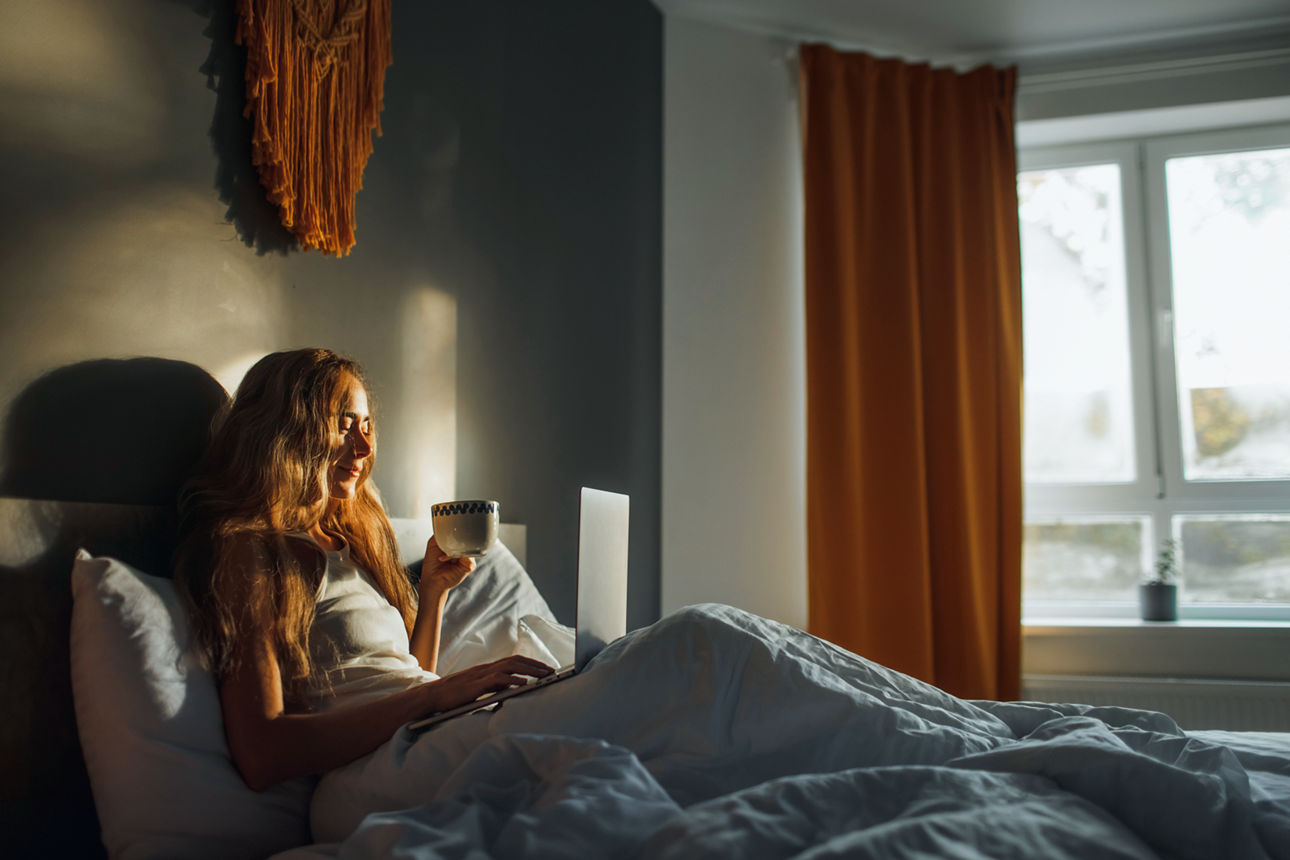 Female relaxation in bedroom, lazy awakening. Lady drinking cappuccino and reading news online on computer. Working from home, freelance. Business and distant job.
