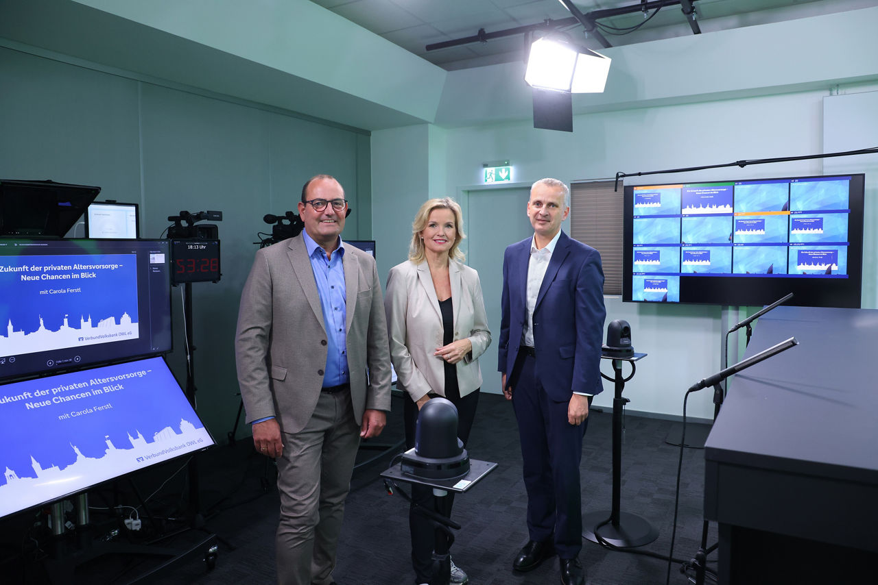 Carola Ferstl steht mit Thorsten Wolff und Andreas Decker im Sendestudio der VerbundVolksbank OWL eG mit Monitoren, Kameratechnik und Beleuchtung.
