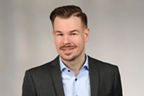 Mann mit braunem, zurückgekämmtem Haar trägt dunklen Anzug und hellblaues Hemd vor neutralem Hintergrund