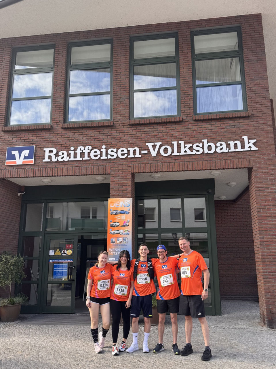 Fünf Personen in orangenen T‑Shirts stehen vor dem Eingang der Raiffeisen‑Volksbank Oder‑Spree eG.