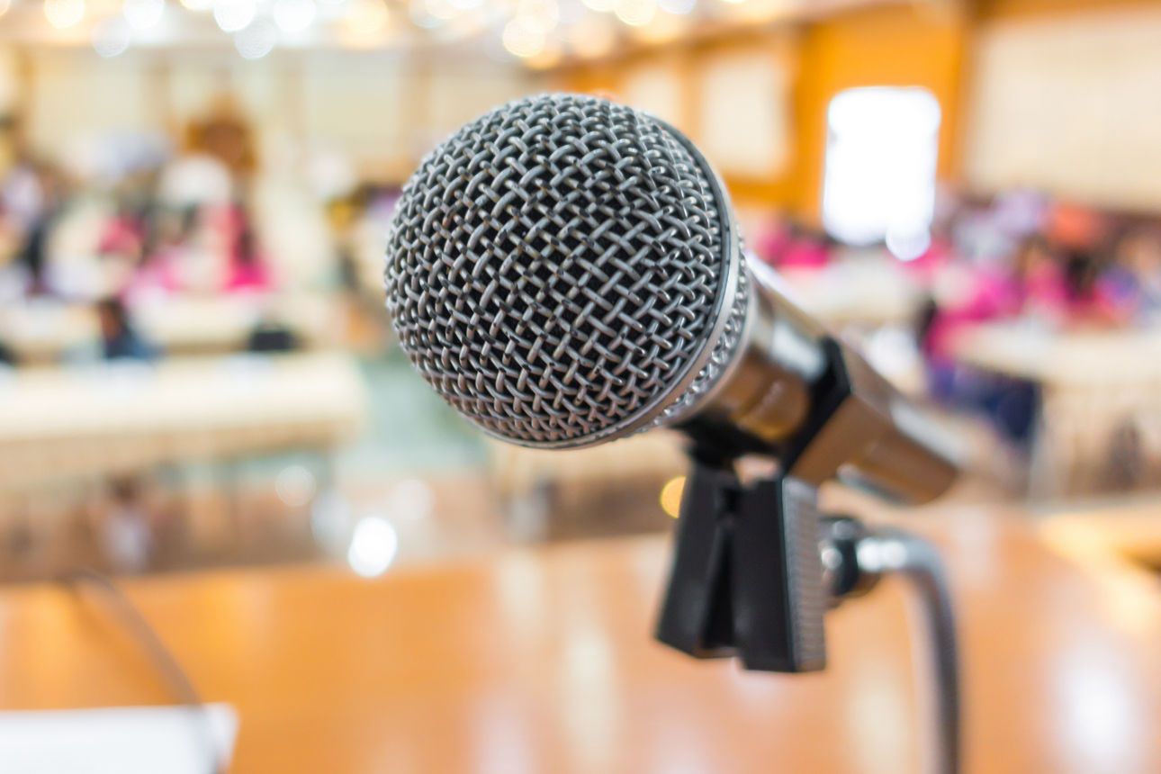 Schwarzes Mikrofon im Vordergrund, dahinter ein unscharfer, heller Konferenzraum.