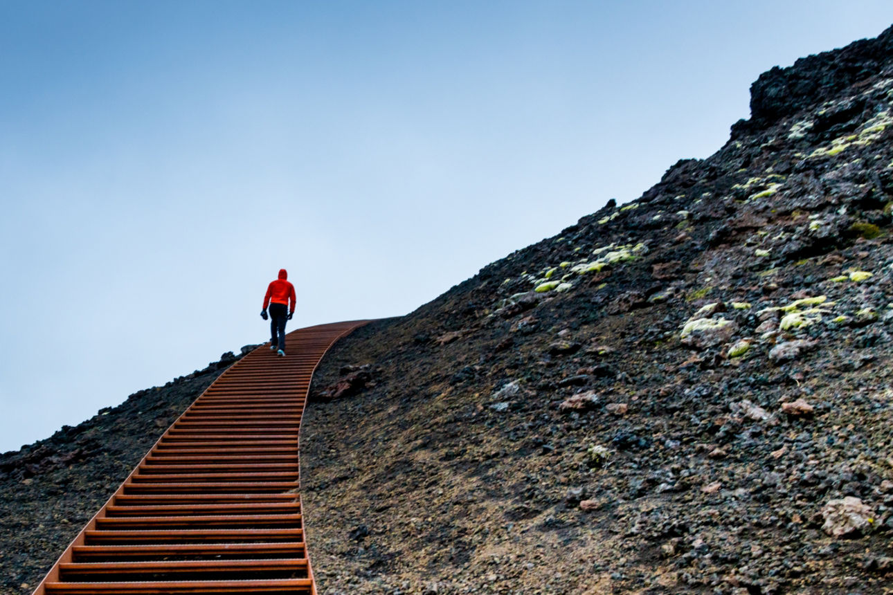Eine einzelne Person in roter Jacke steigt eine lange Treppe auf einem steinigen Hang unter blauem Himmel hinauf