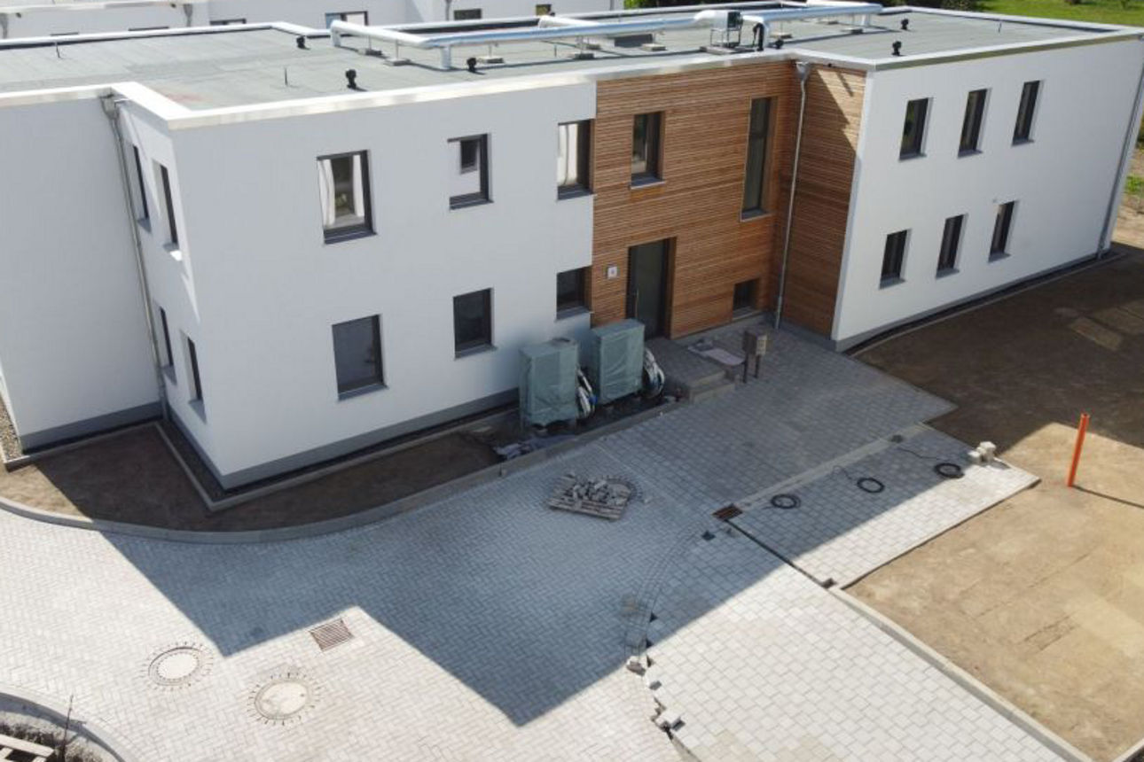 Zweigeschossiges, modernes Gebäude mit weißer Fassade und Holzelementen, umgeben von gepflasterten Wegen und einer offenen La