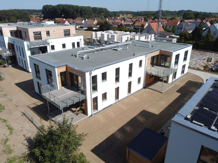 Luftaufnahme mehrerer moderner Flachdachgebäude in einer Neubausiedlung, umgeben von Feldern und Einfamilienhäusern.