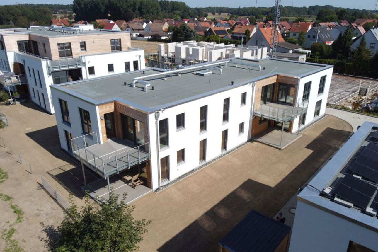 Luftaufnahme mehrerer moderner Flachdachgebäude in einer Neubausiedlung, umgeben von Feldern und Einfamilienhäusern.