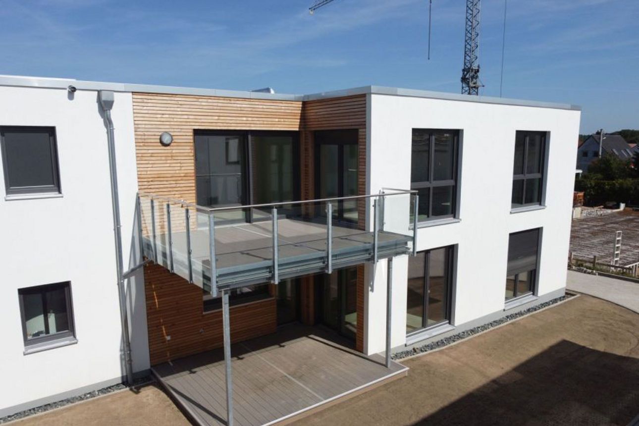 Zweigeschossiges Gebäude mit weißer Fassade, Holzelementen und einem großen Balkon mit Glasgeländer.