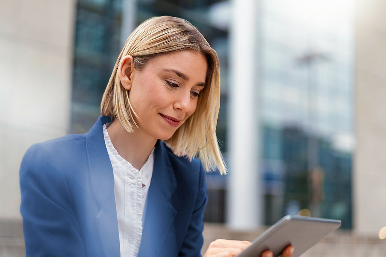 Junge, blonde Geschäftsfrau mit weißer Bluse und blauem Blazer, tippt und schaut auf ihr Tablet. Im Hintegrund sieht man ein Geschäftsgebäude in absoluter Unschärfe.