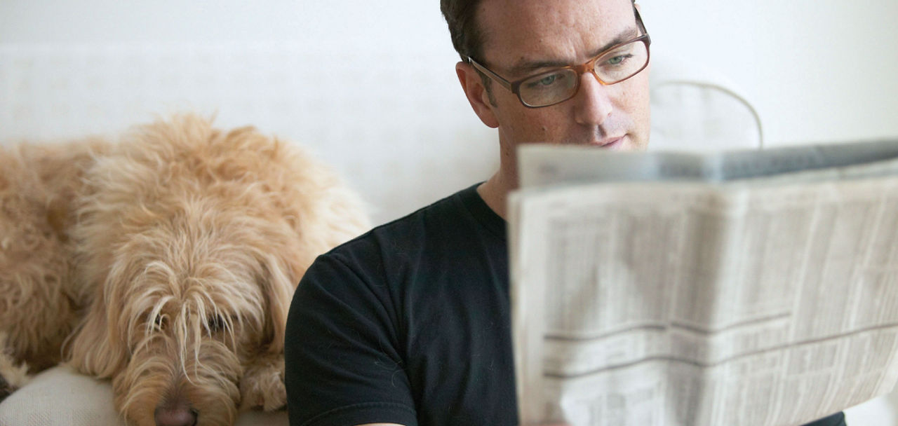 Ein Mann sitzt auf dem Sofa und liest eine Zeitung. Neben ihm liegt ein Hund.