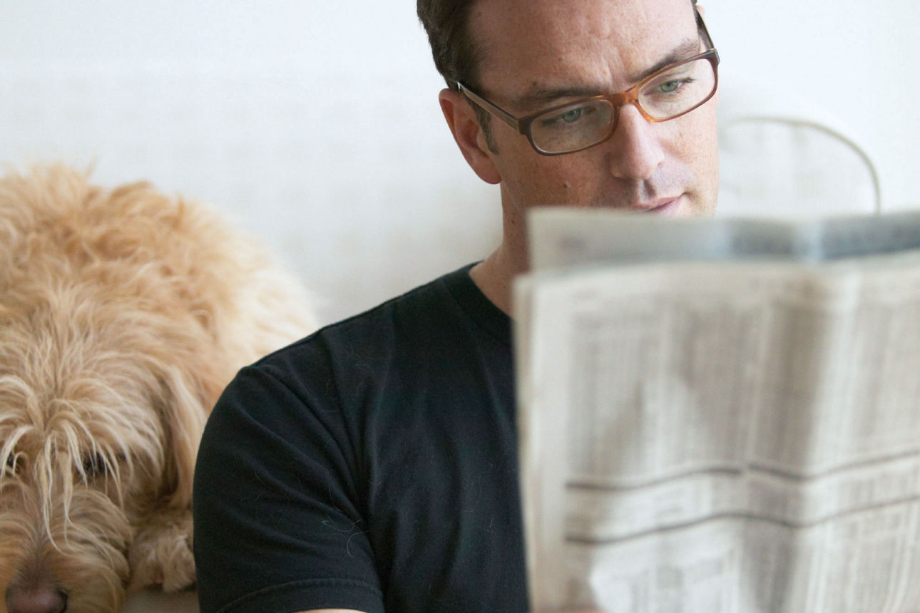 Ein Mann sitzt auf dem Sofa und liest eine Zeitung. Neben ihm liegt ein Hund.