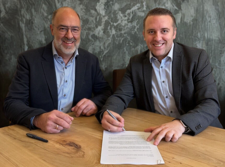 Zwei Herren sitzen zu ener Vertragsunterzeichnung an einem Tisch und lächeln in die Kamera