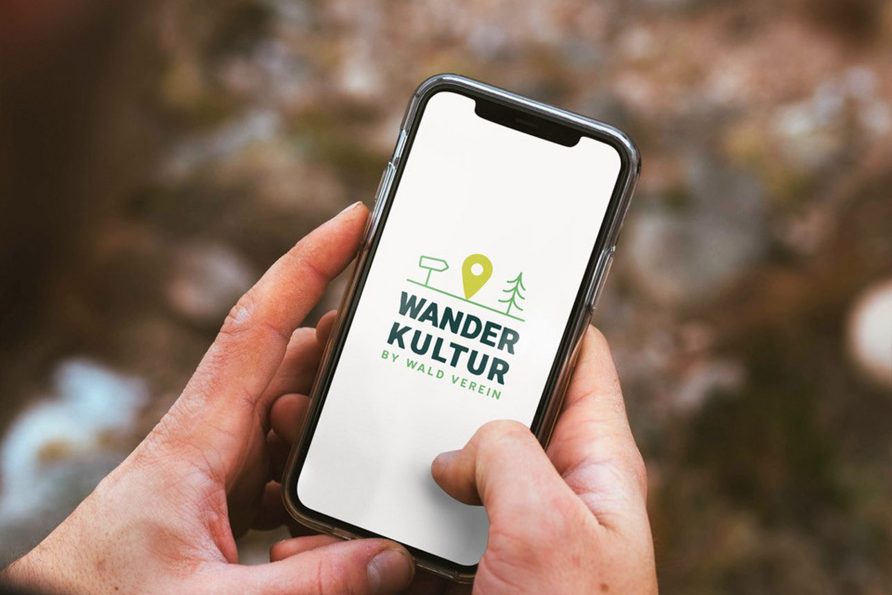 Hände halten ein Smartphone mit dem Logo „Wander Kultur“ vor einem unscharfen, natürlichen Hintergrund.