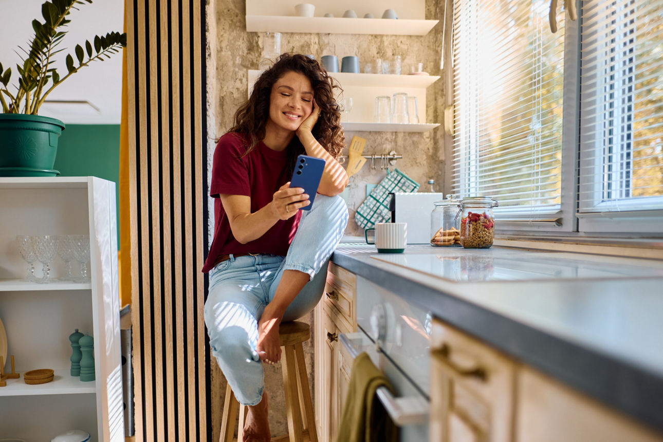 junge Frau sitzt in ihrer Küche und schaut lächelnd auf ihr Smartphone