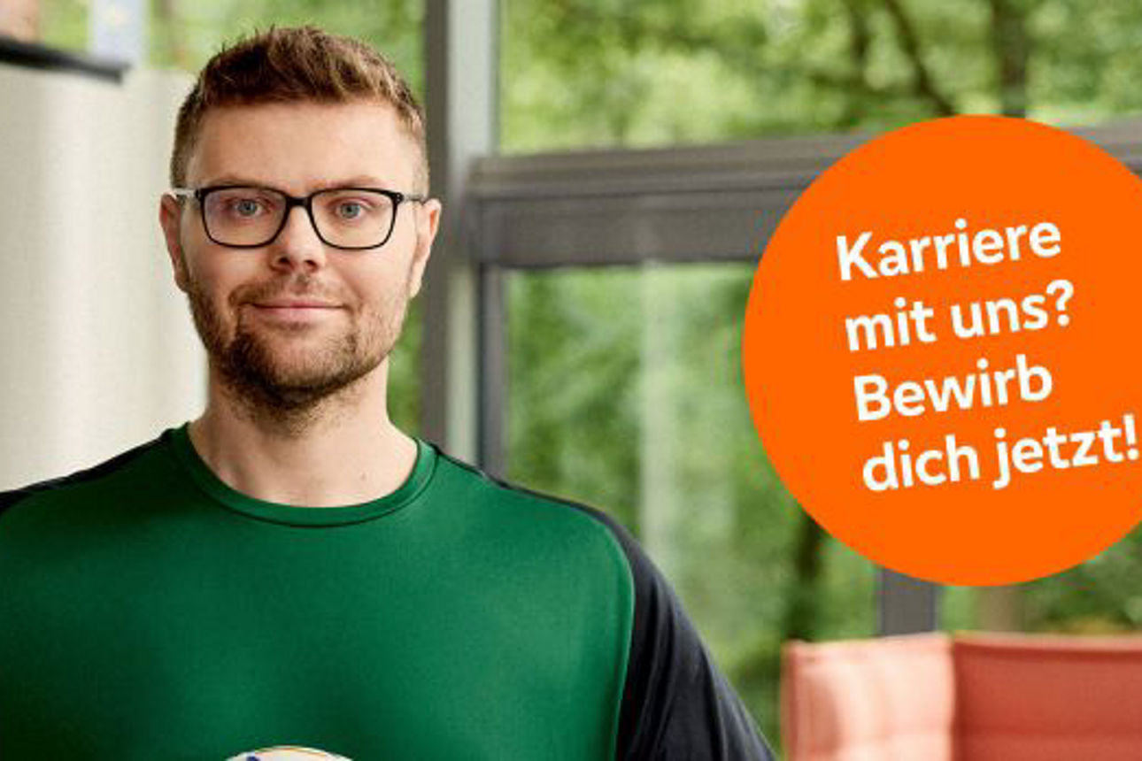Ein junger Mann in einem grünen Sportshirt steht vor einer Fensterfront und hält einen Handball