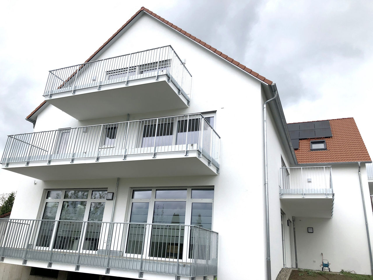 Das Bild zeigt ein modernes Haus mit drei Stockwerken. Die Fassade ist weiß. Das Dach ist rot und hat Ziegel. Auf allen Etagen gibt es große Fenster. Unten im Erdgeschoss ist eine überdachte Veranda. Sie wird von mehreren Säulen gehalten. Vor dem Haus ist ein gepflasterter Weg. Rechts führt eine Rampe zu einer Garage oder einem Keller.