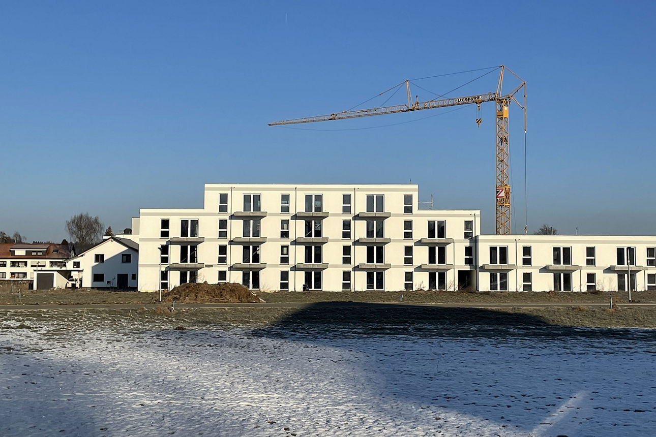 Weißes Neubau Mehrfamilienhaus. Davor eine teilweise schneebedeckte Wiese. Der Himmel ist blau. Es ist ein Kran zu sehen.