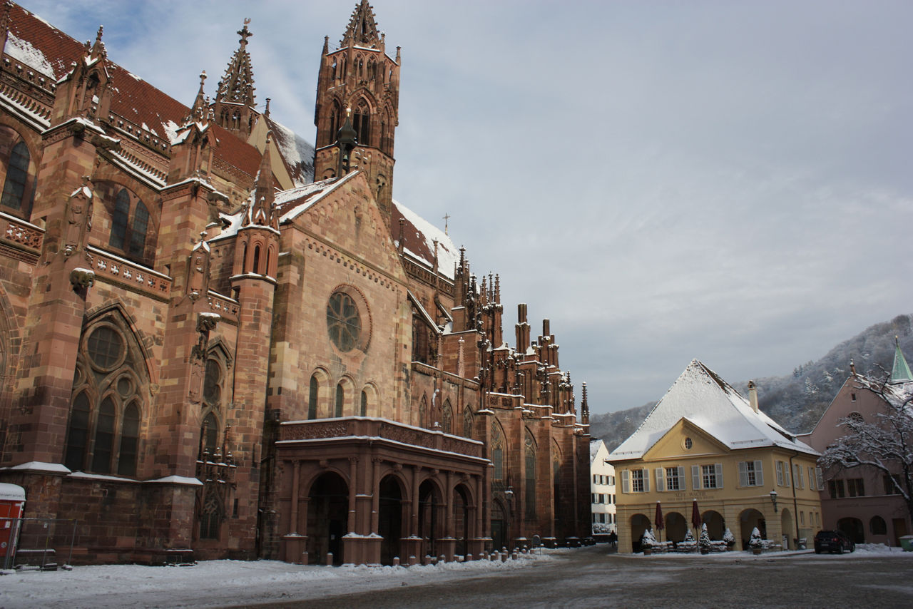 Das Bild zeigt eine verschneite Altstadt mit einer großen gotischen Kirche aus rotem Sandstein links und einem gelben historischen Gebäude mit Arkaden rechts auf einem winterlichen Platz vor bewaldeten Hügeln.