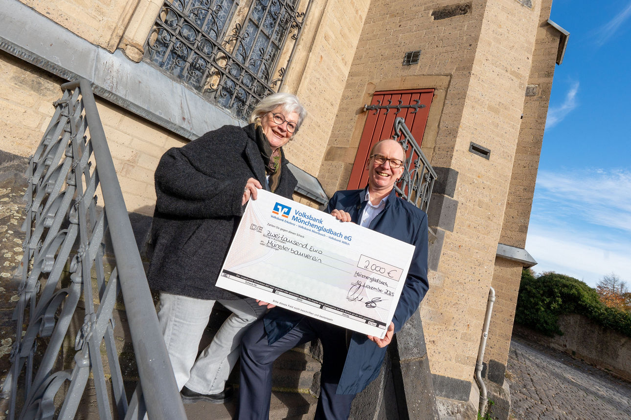 Auf dem Foto stehen zwei Personen auf einer Steintreppe vor einem historischen Kirchengebäude und halten einen Scheck.