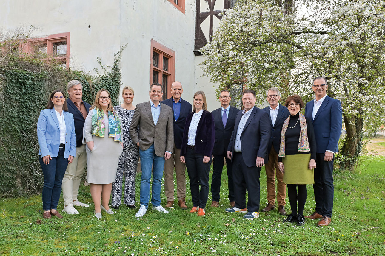 5 Frauen und 7 Männer stehen auf einer Wiese für einem alten Gebäude und lächeln in die Kamera