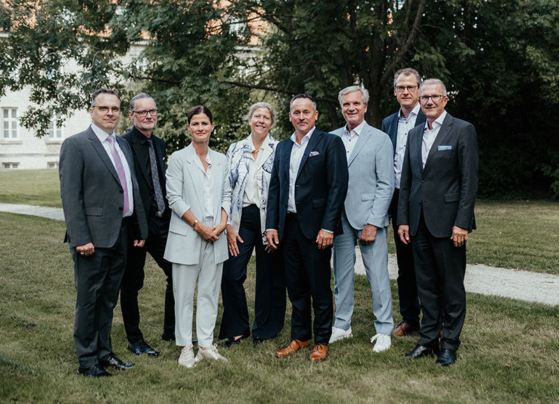 Die acht Mitgleider des Aufsichtsrats stehen als Gruppe aufgestellt in einem Garten. 