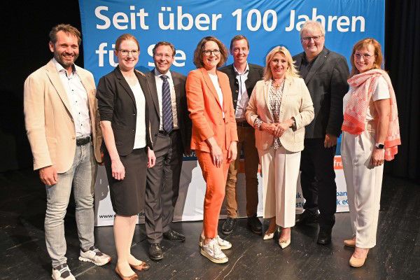 Eine Gruppe von sieben Personen steht vor einem blau-weißen Transparent.