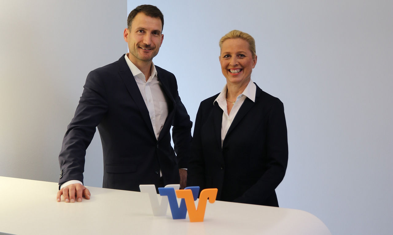 Sabine Haas und Christoph Appel im Business-Outfit vor hellem Hintergrund, lächelnd am weißen Tresen mit VR-Logo.