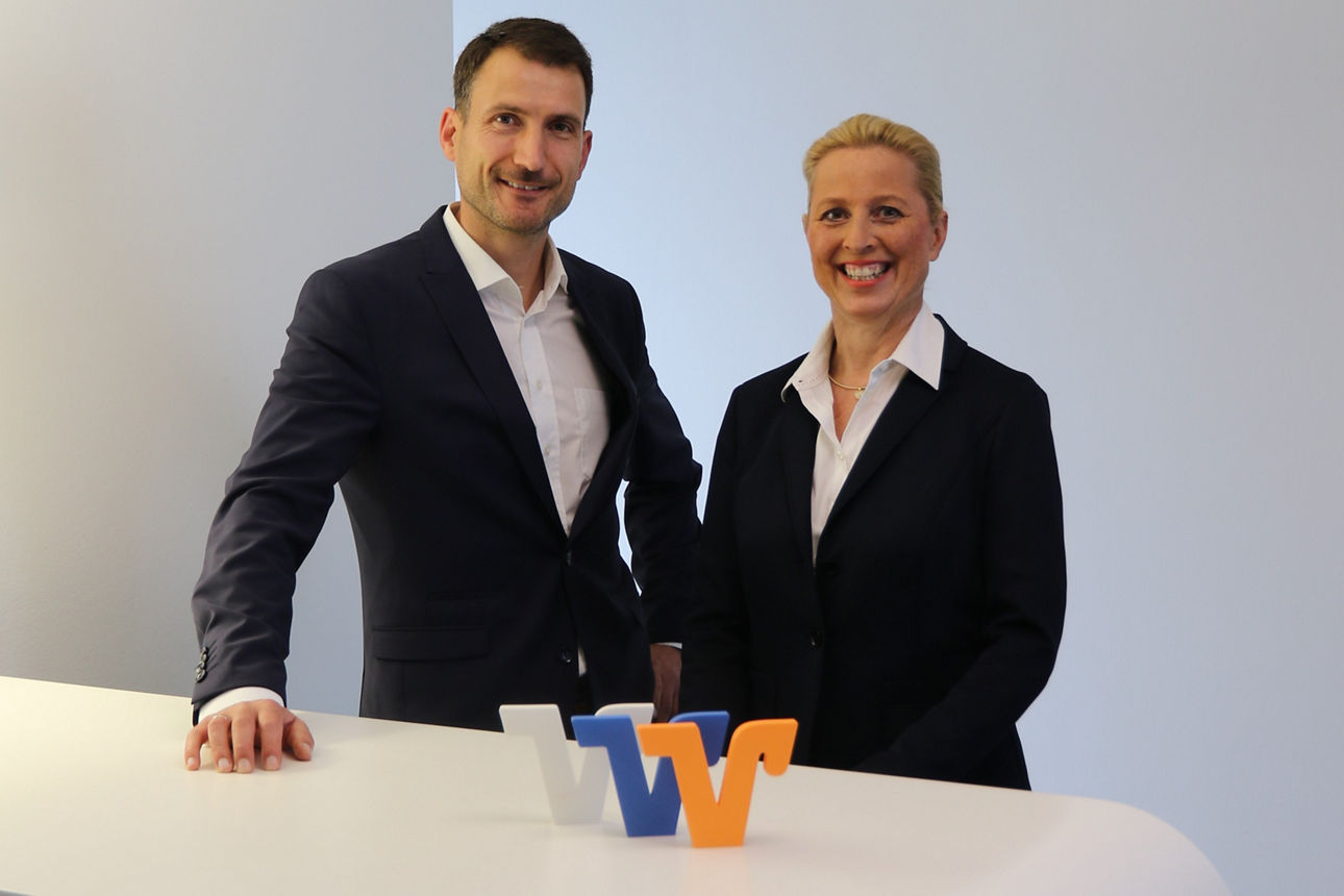 Sabine Haas und Christoph Appel im Business-Outfit vor hellem Hintergrund, lächelnd am weißen Tresen mit VR-Logo.