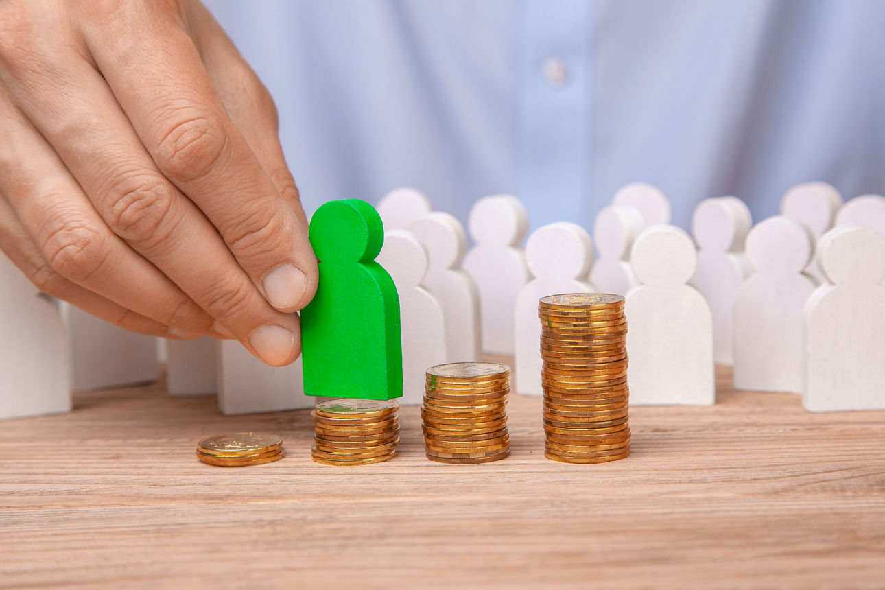 Eine Hand hebt eine Holzfigur von einem Münzhaufen auf einen höheren.