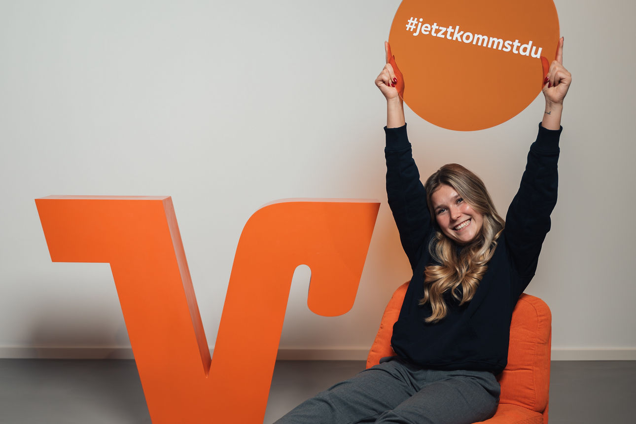 Junge Person auf orangefarbenem Stuhl hält Schild mit #jetztkommstdu vor großem VR-Logo der VR Bank RheinAhrEifel eG.