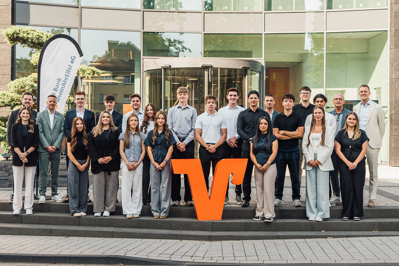 Mitarbeitende der VR Bank RheinAhrEifel eG vor modernem Bankgebäude, gruppiert um großes oranges V-Logo