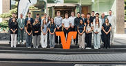 Mitarbeitende der VR Bank RheinAhrEifel eG vor modernem Bankgebäude, gruppiert um großes oranges V-Logo
